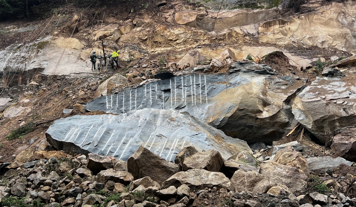 Drilling crews install steel dowels into a fractured rock face at the I-5 landslide site south of Bellingham after crews split a large boulder; debris remains below the slope.